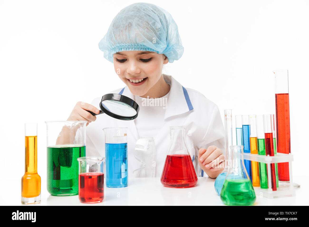 Portrait of cheerful little girl in white laboratory coat holding magnifying glass during chemical experiments isolated over white background Stock Photo