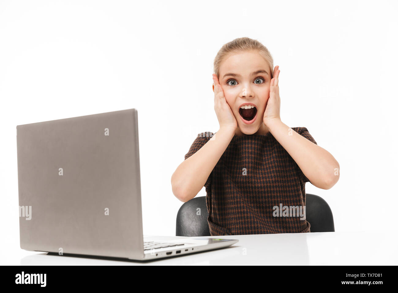 Portrait of shocked school girl rejoicing and using silver laptop while sitting at desk in class isolated over white background Stock Photo