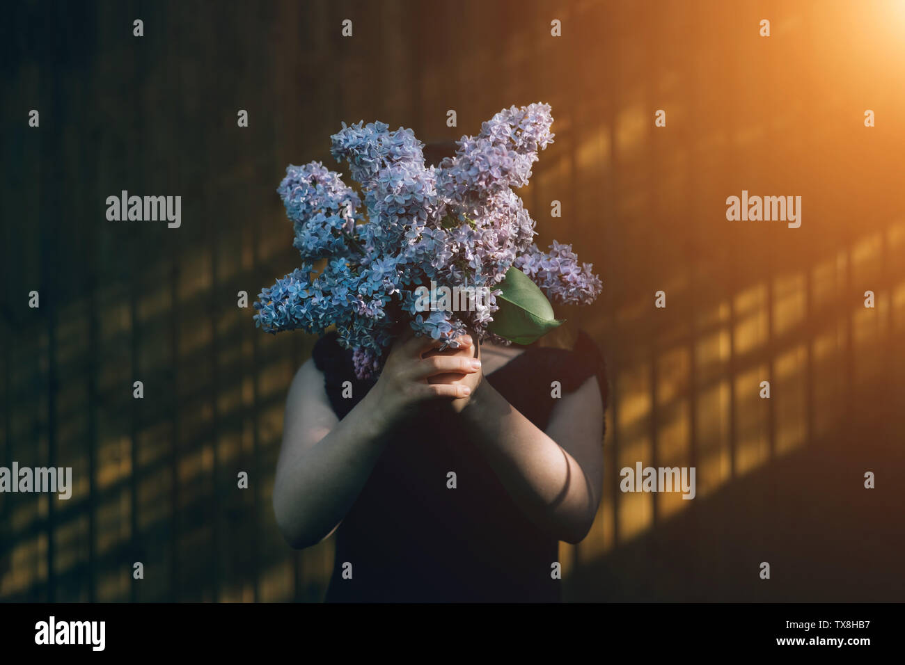 Girl holding lilac bouquet in front of face. Child enjoying flowers outdoors in front of wall. Kid hiding behind bunch of violet flowers. Summer and spring flora. Young person with gift in hands Stock Photo