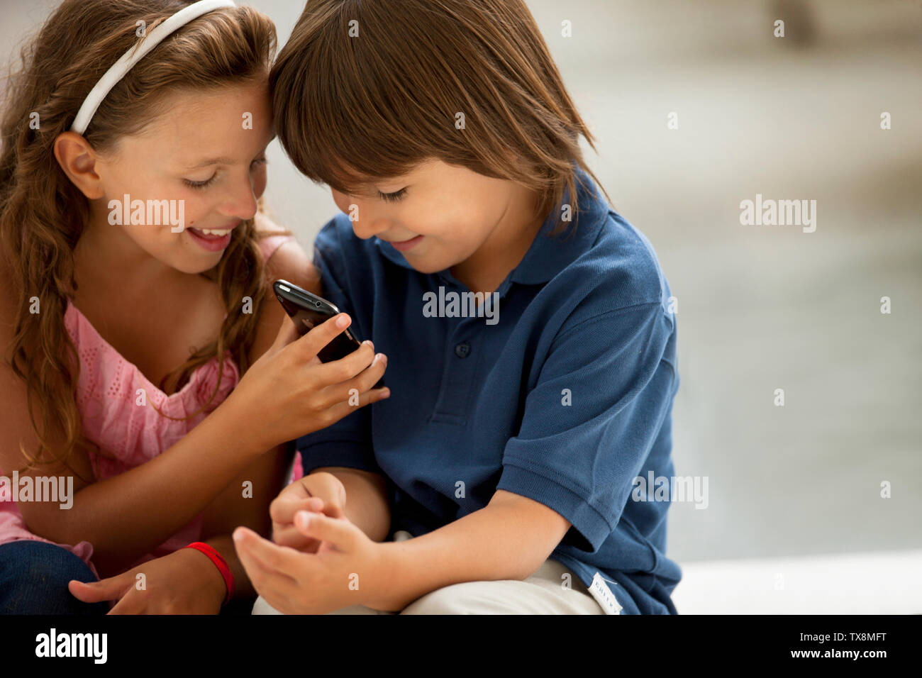 A young boy and girl smile mischievously as they look at a cellphone. Stock Photo