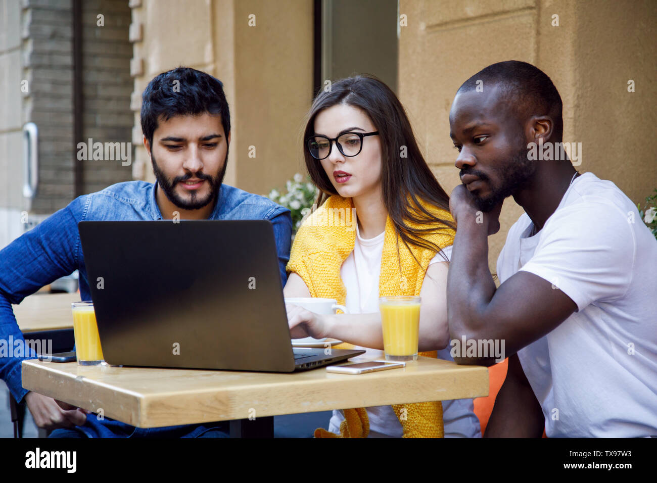 Three multicultural smart students discussing something at the laptop while studying at the cafe Stock Photo
