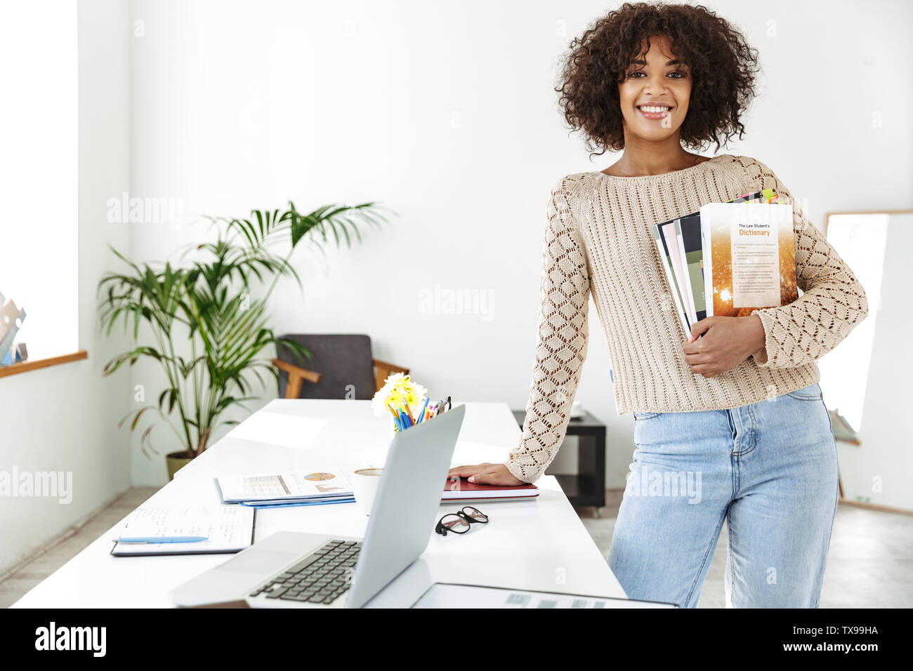 Cheerful african woman wearing in casual clothes holding magazines and looking at the camera while standing near the table at office Stock Photo