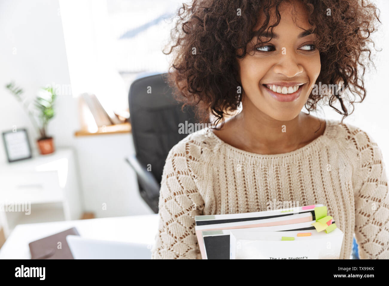 Smiling african woman wearing in casual clothes holding magazines and looking away while standing at office Stock Photo