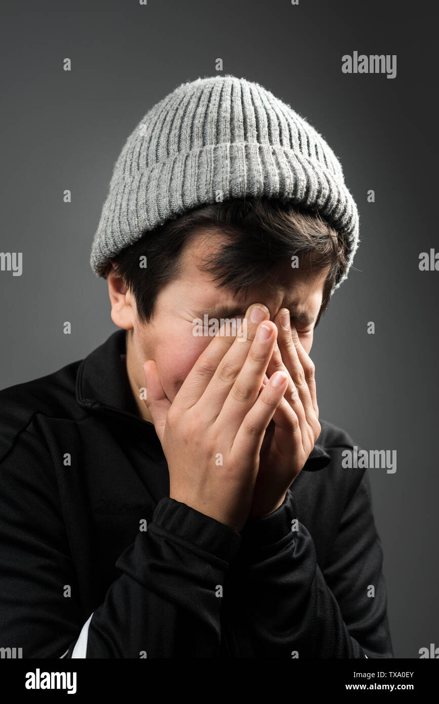 Young boy, 11 years old in black tracksuit and grey wooly hat crying with his face in his hands Stock Photo