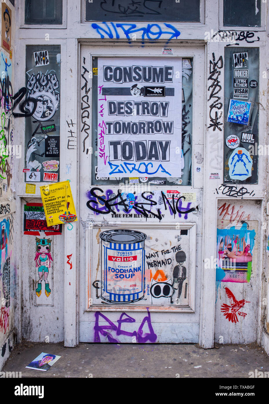 Door covered in graffiti and stickers with poster from environmental campaign saying Consume destroy tomorrow today in Shoreditch, London Stock Photo