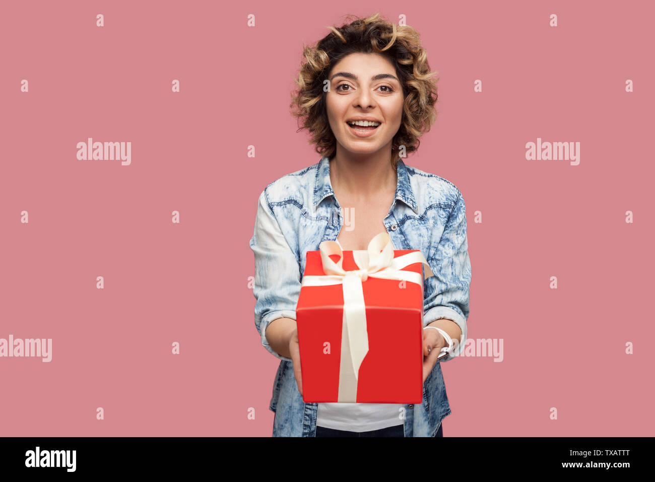 Portrait of happy beautiful young woman in blue shirt with curlty hairstyle standing and giving you red gift box with toothy smile, looking at camera. Stock Photo
