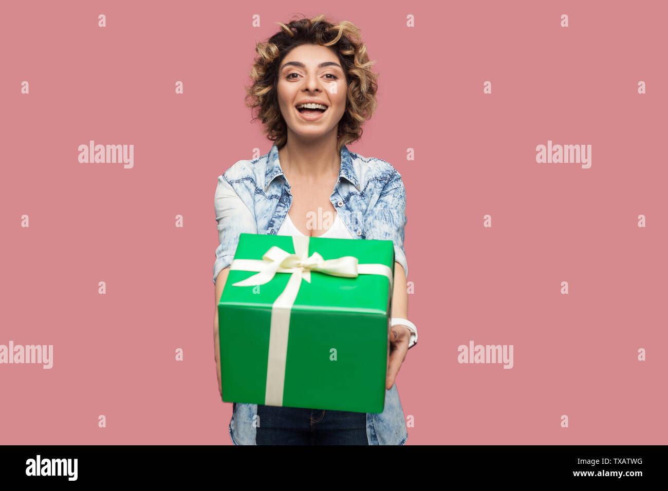 Portrait of happy beautiful young woman in blue shirt with curlty hairstyle standing and giving you green gift box with toothy smile, looking at camer Stock Photo