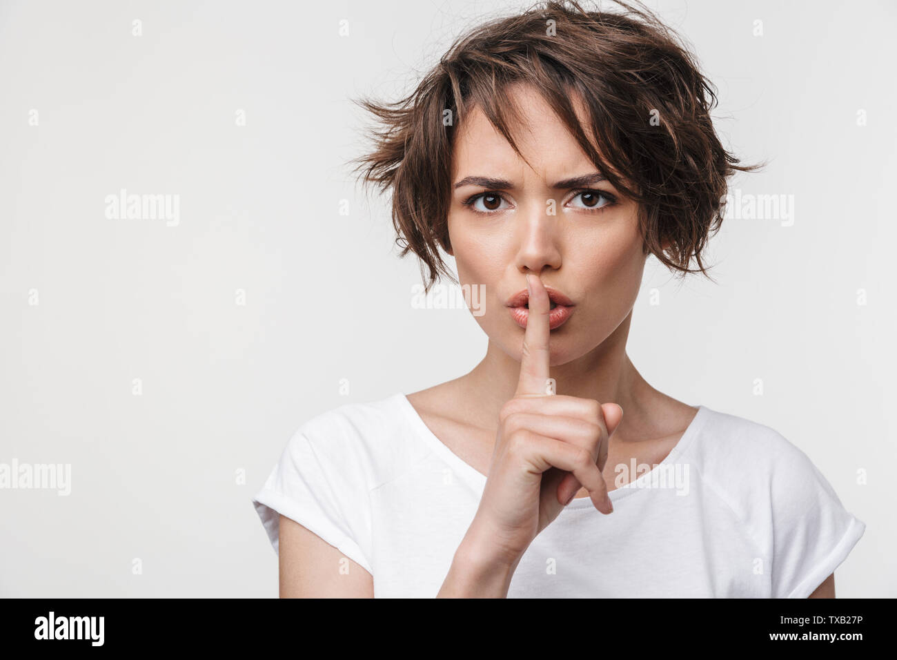 Portrait of strict woman with short brown hair in basic t-shirt holding index finger on lips while standing isolated over white background Stock Photo