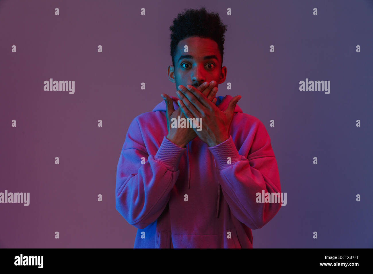 Portrait of surprised african american man in colorful hoodie poising at camera with hands on his mouth isolated over violet background Stock Photo