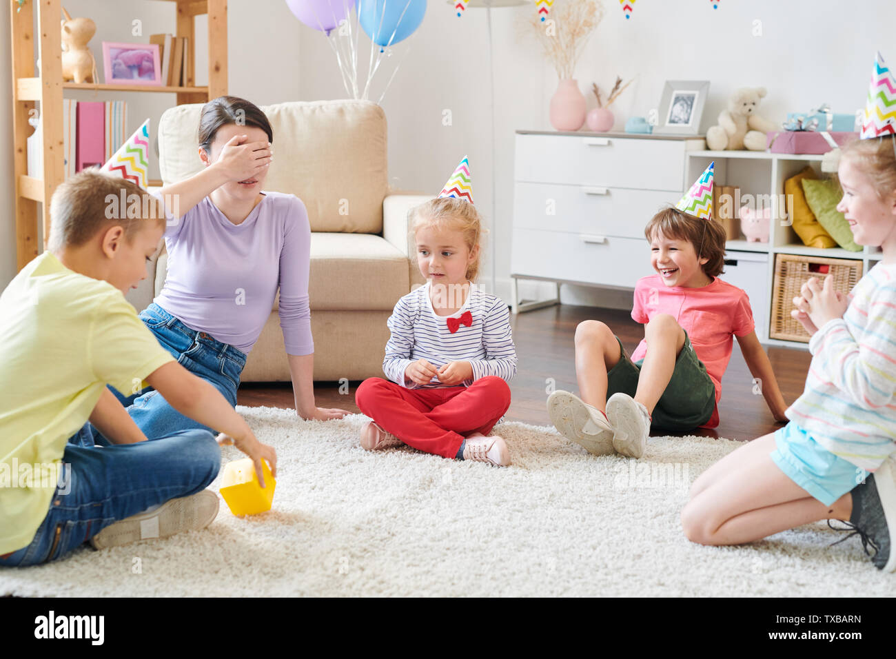 Group of cheerful little kids in birthday caps sitting in circle on rug Stock Photo