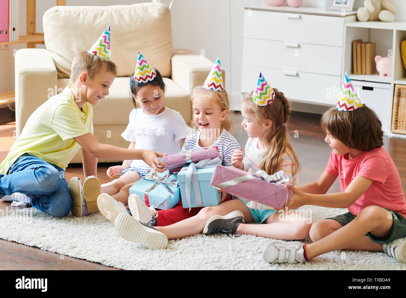 Group of adorable friends in birthday caps giving presents to happy blonde girl Stock Photo
