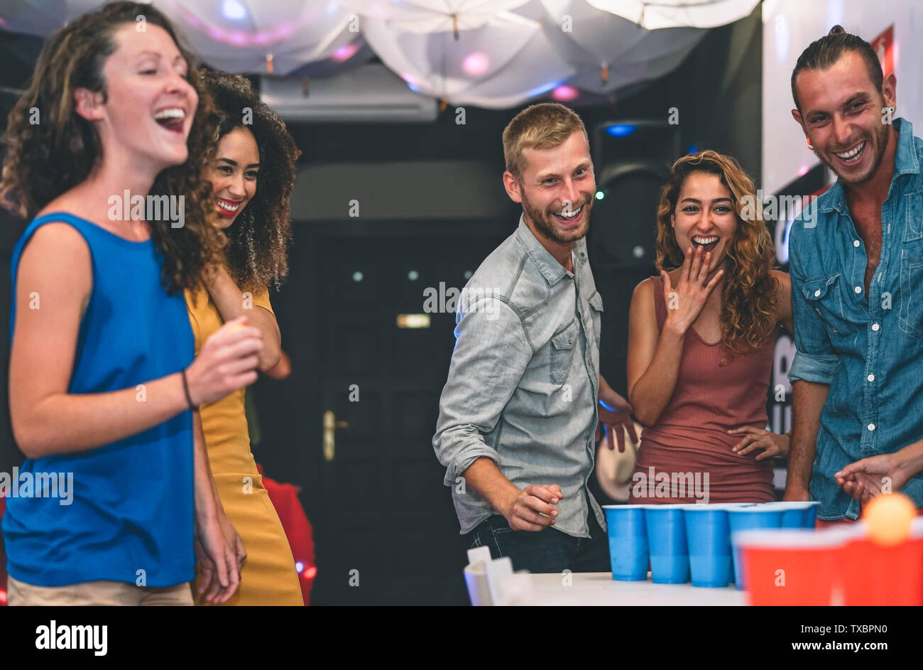 Happy friends playing beer pong in a cocktail bar - Young millennials people having fun doing party alcohol games at night pub Stock Photo