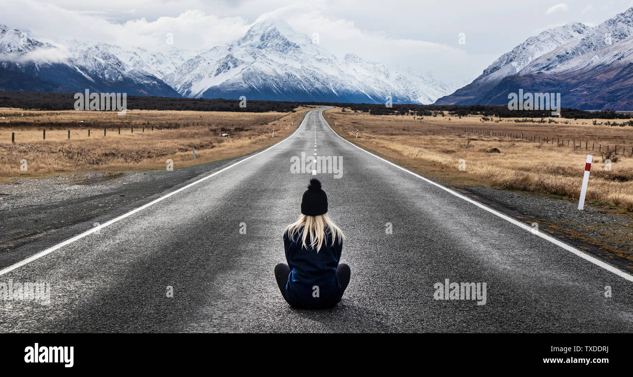 Girl with blonde hair wearing a hat sits in the middle of the road in New Zealand looking at the mountain view Stock Photo