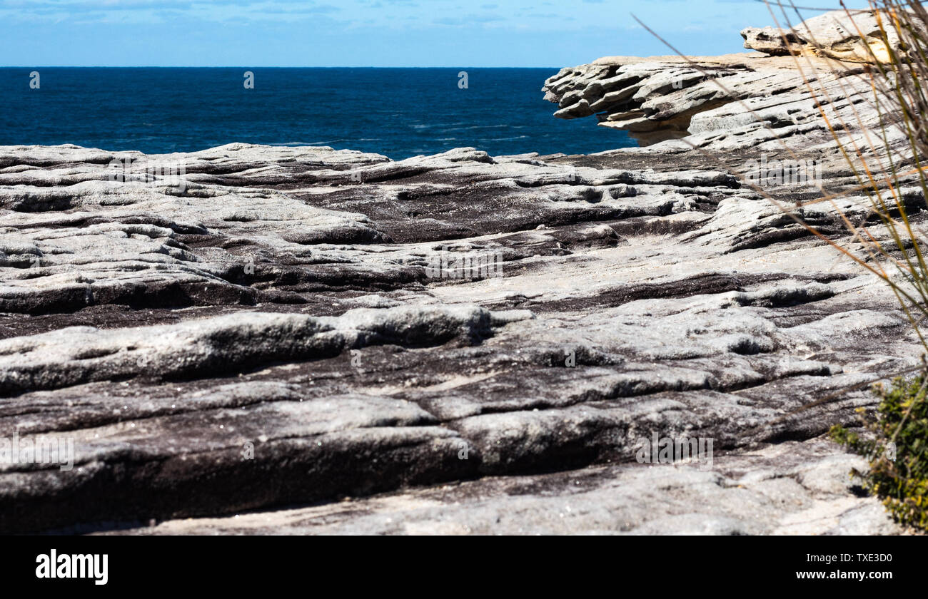 Oceanside rocky cliff top sandstone wave ridges with blue coastal sea and sky in background Stock Photo
