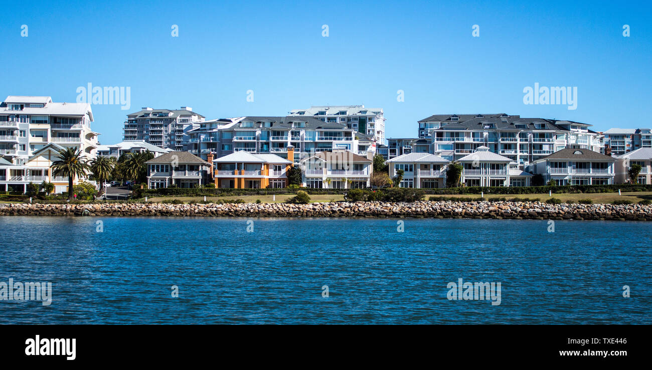 Harbourside condominium apartments behind free standing houses with grass frontage, stone retaining wall and blue river against blue sky Stock Photo