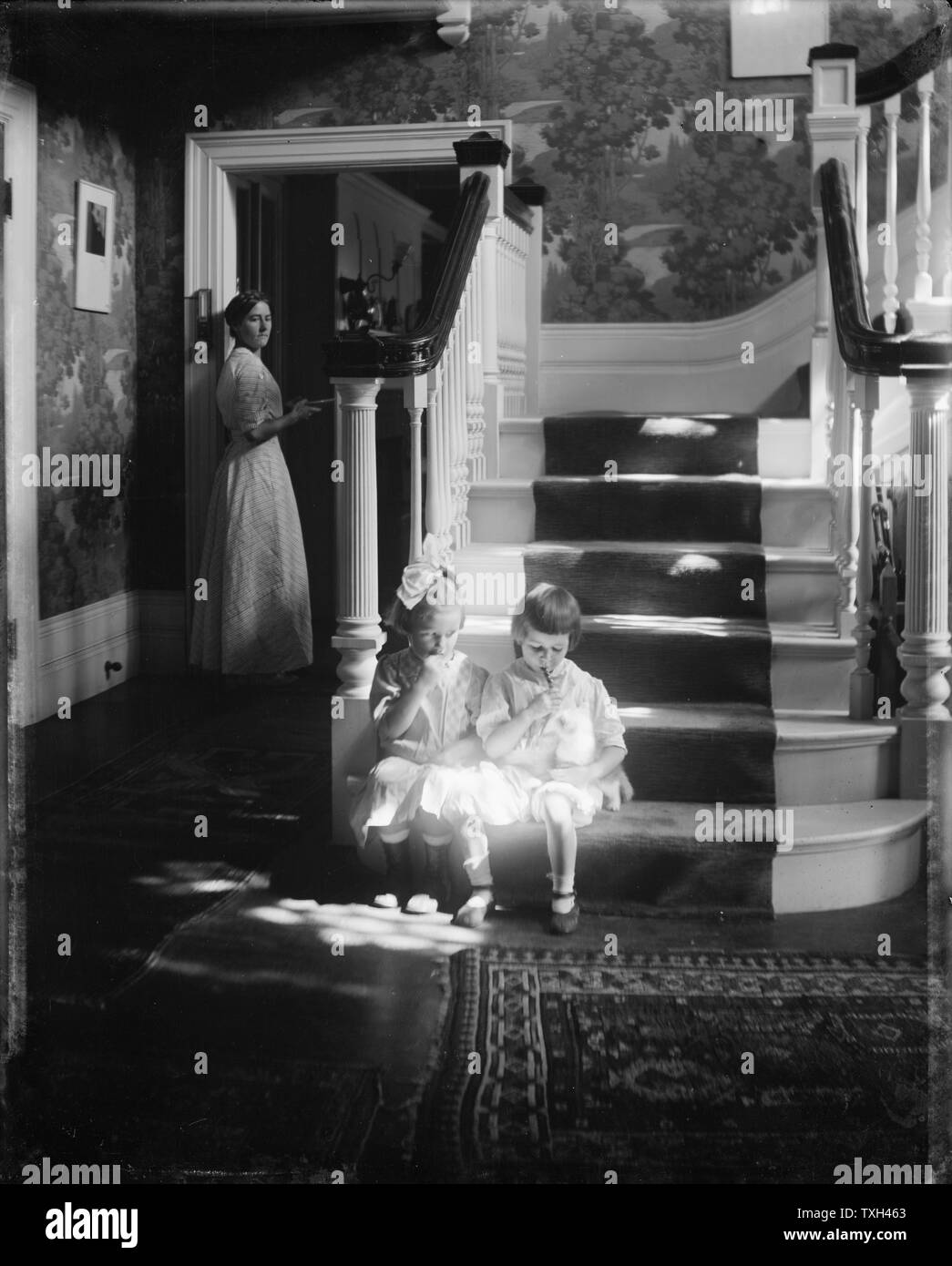 Gertrude Käsebier American school Lollipops : Photograph of two small girls sitting at the bottom of the stairs in a shaft of sunlight, sucking lollipops, while a woman watches from a doorway. Stock Photo