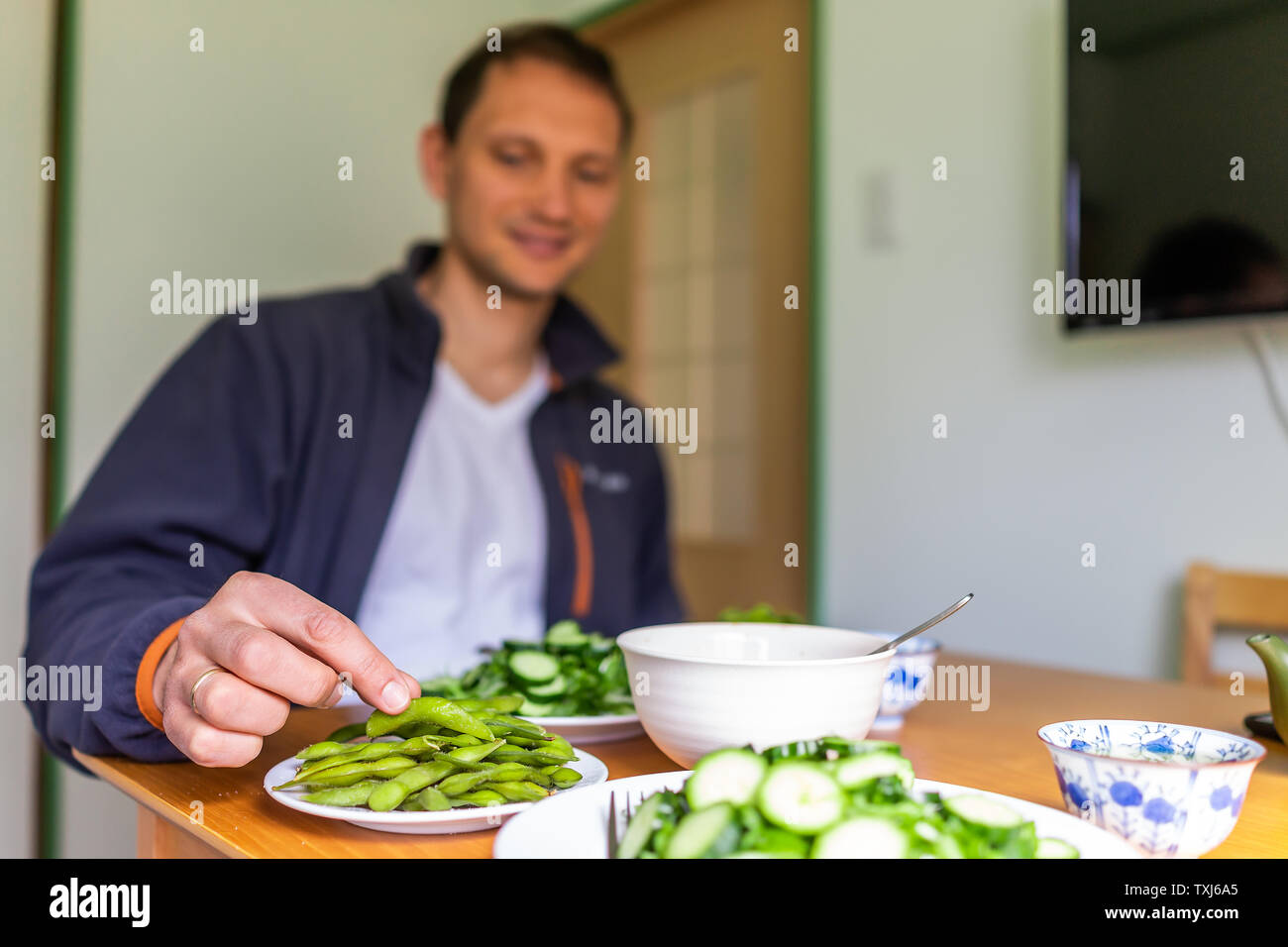 Home with table and green salad dish with Japanese cucumbers and mizuna greens and man trying boiled edamame soy beans Stock Photo