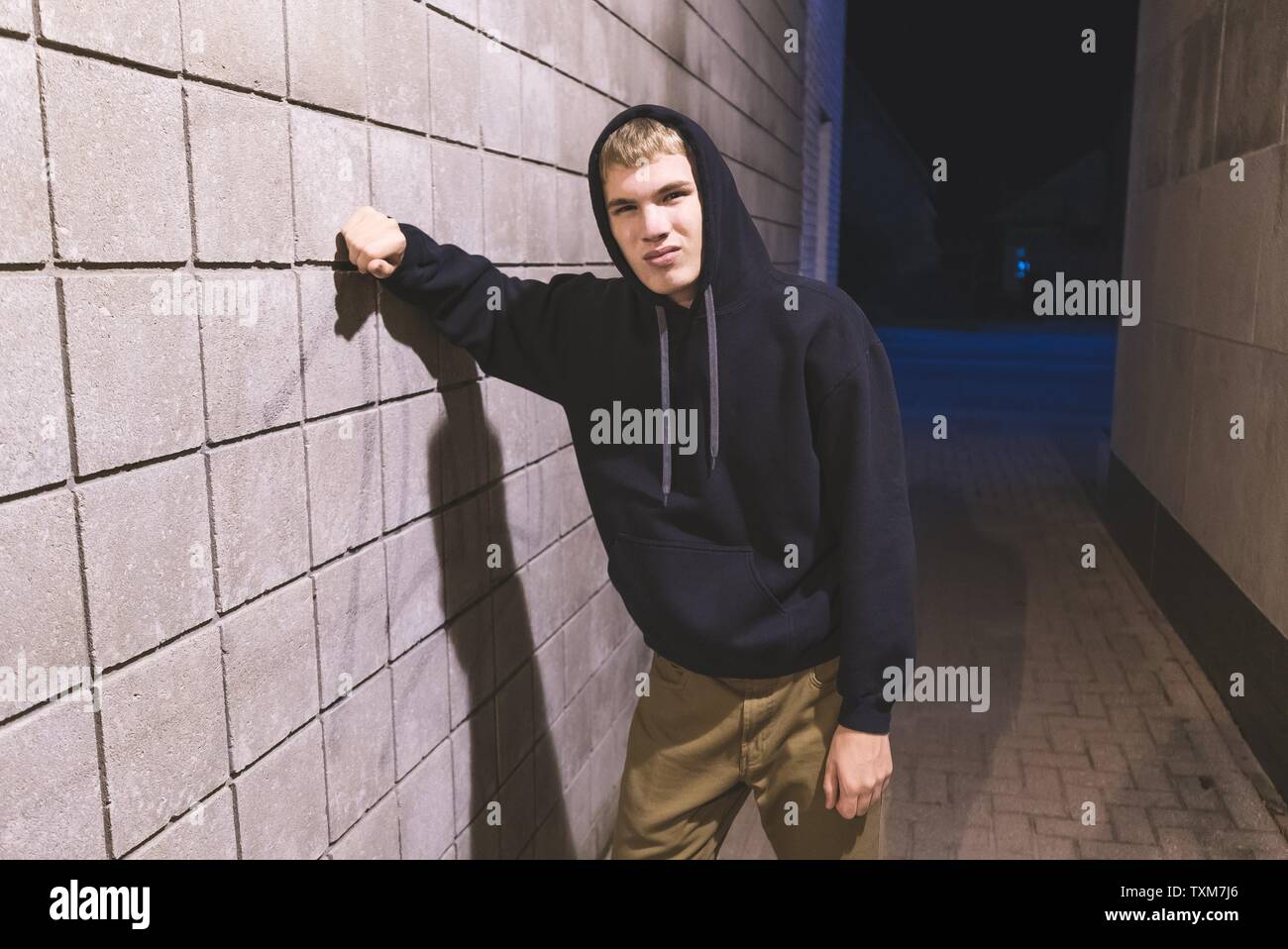 Hooded teenager leaning against a brick wall in an alleyway. He is ...