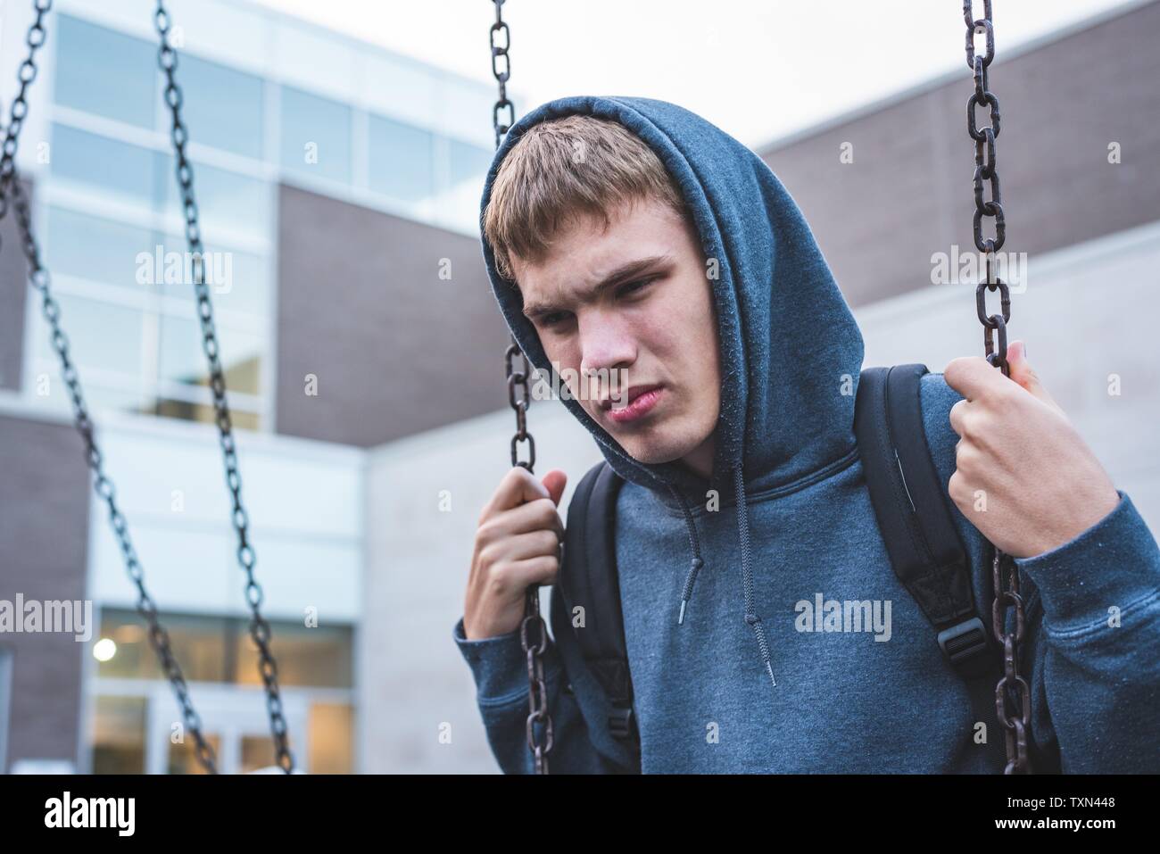 Sad teenager sitting on a swing outside of a school. He is reminiscing about when he was younger. Stock Photo