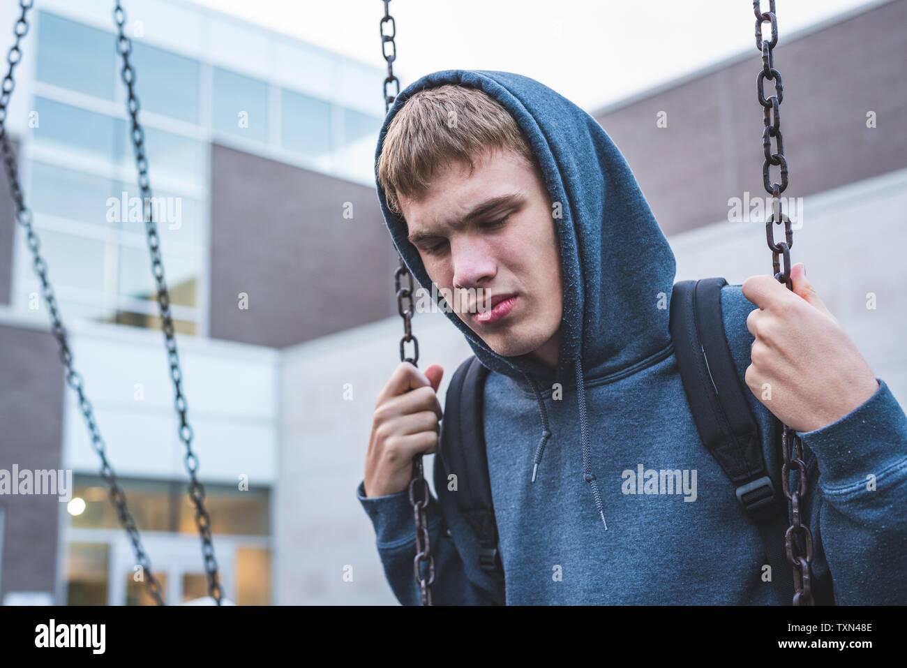 Sad teenager sitting on a swing outside of a school. He is reminiscing about when he was younger. Stock Photo