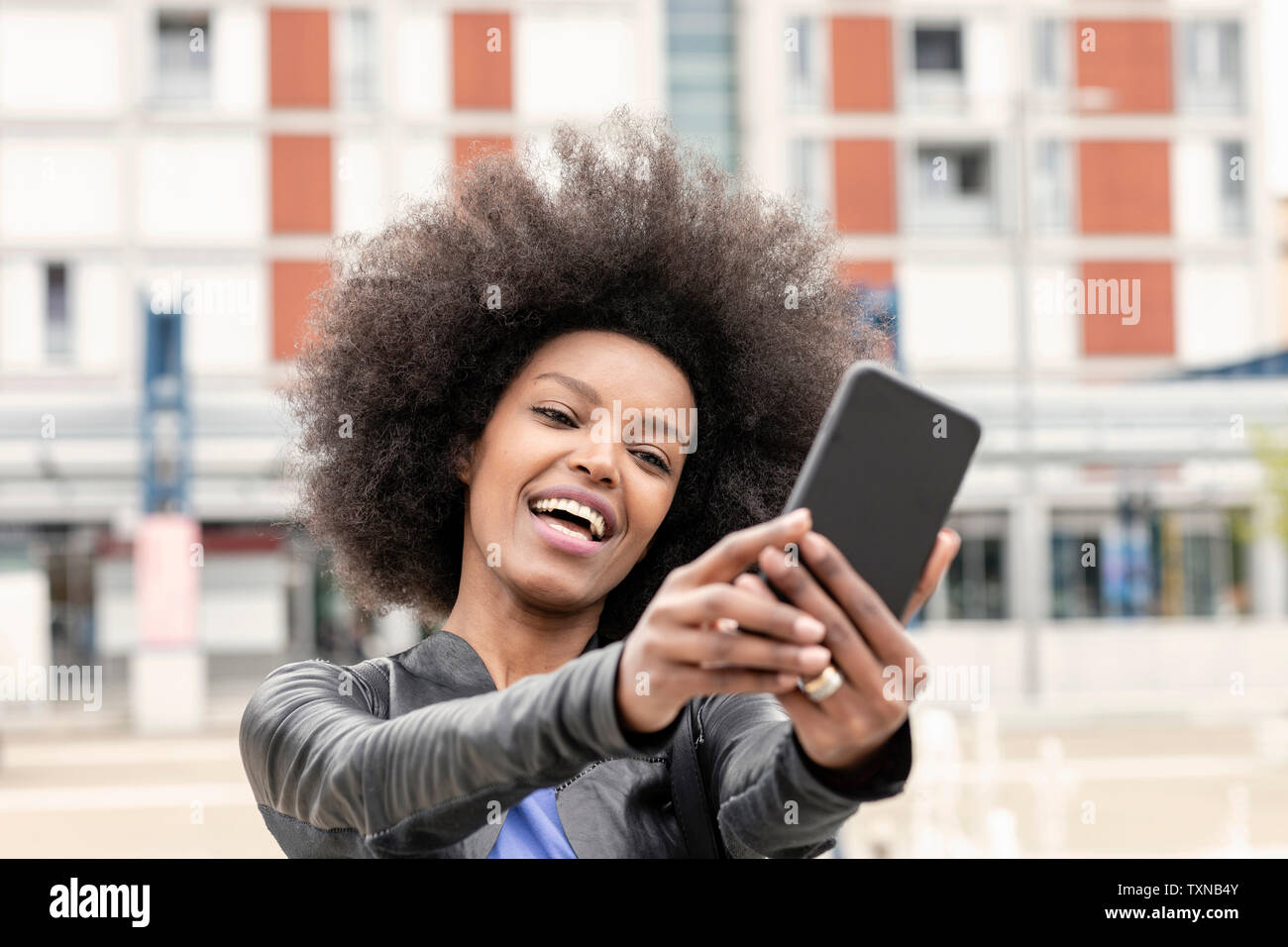Happy young woman with afro hair in city, taking smartphone selfie Stock Photo