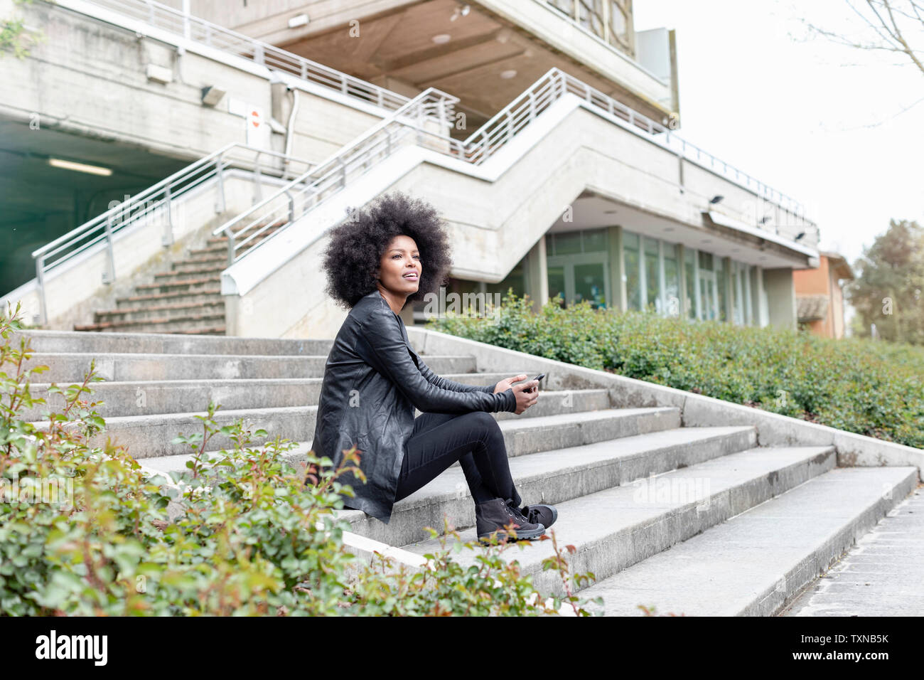 Young woman with afro hair sitting on city stairway looking away Stock Photo