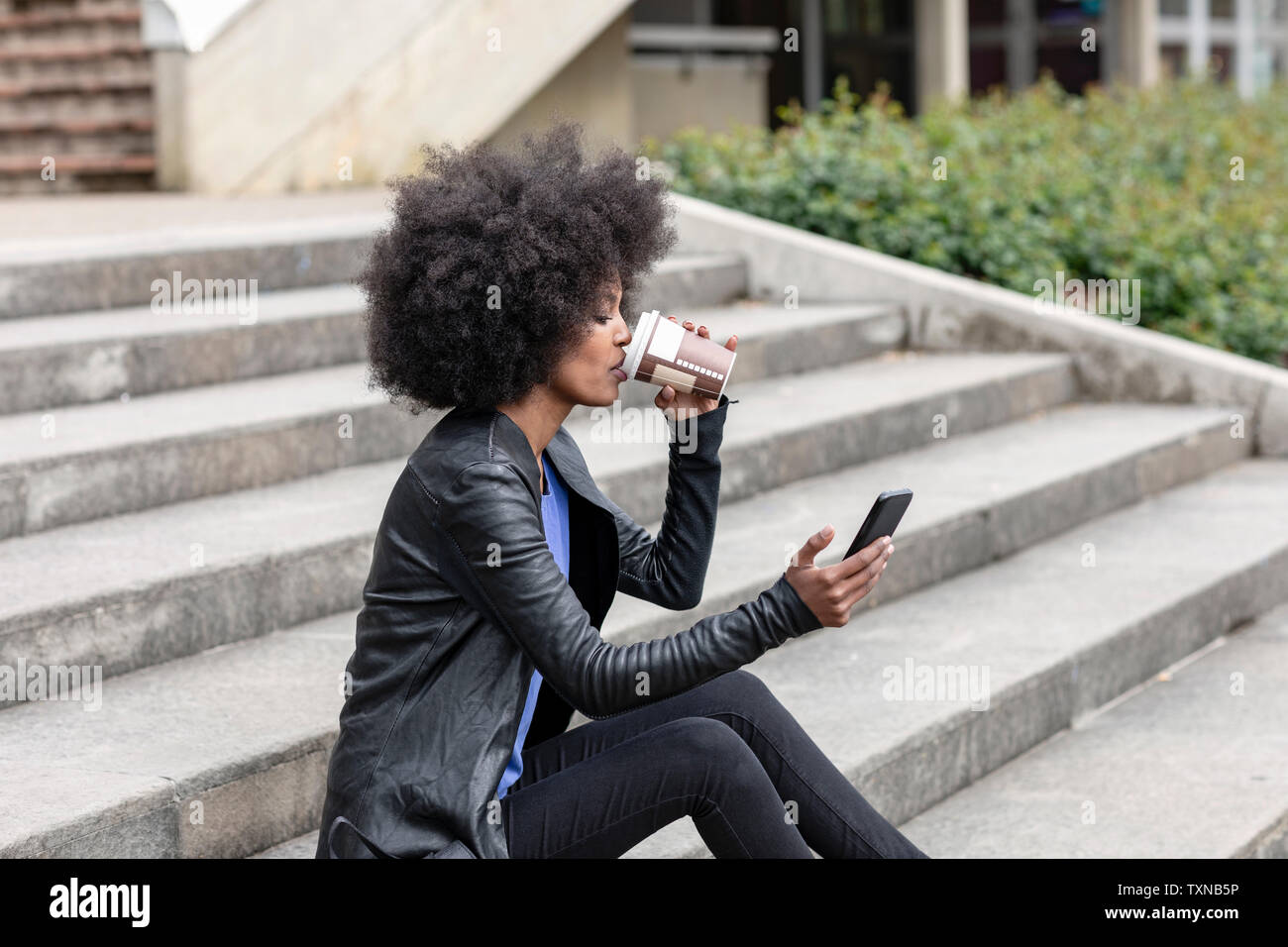 Young woman with afro hair sitting on city stairway, drinking takeaway coffee and looking at smartphone Stock Photo