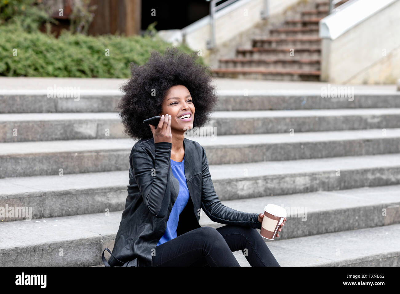 Happy young woman with afro hair sitting on city stairway, listening to smartphone Stock Photo