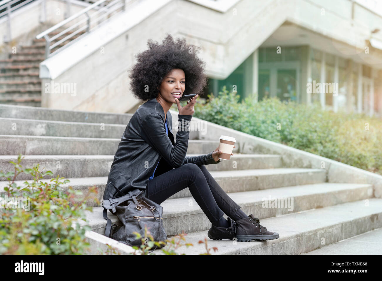 Young woman with afro hair sitting on city stairway, talking to smartphone Stock Photo