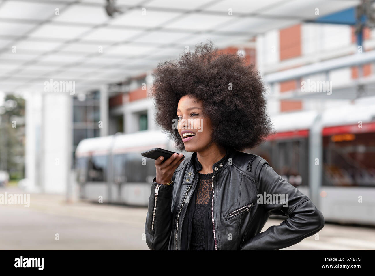 Young woman with afro hair at city train station, talking to smartphone Stock Photo