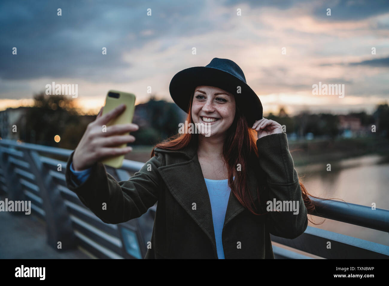 Young woman with long red hair on footbridge taking smartphone selfie at dusk, Florence, Tuscany, Italy Stock Photo