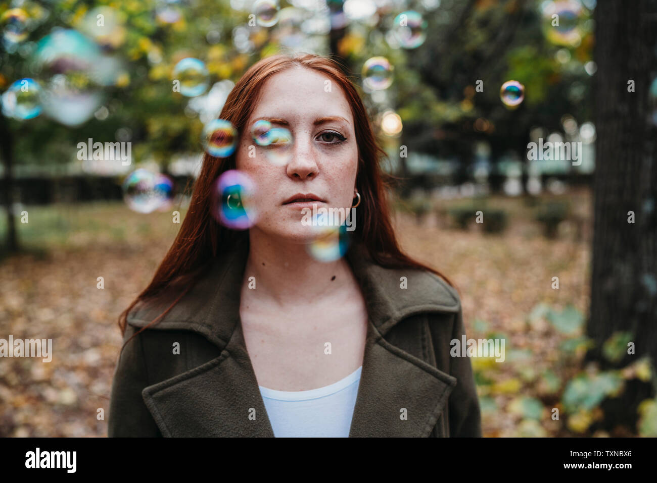 Young woman with long red hair amongst floating bubbles in autumn park, shallow focus portrait Stock Photo