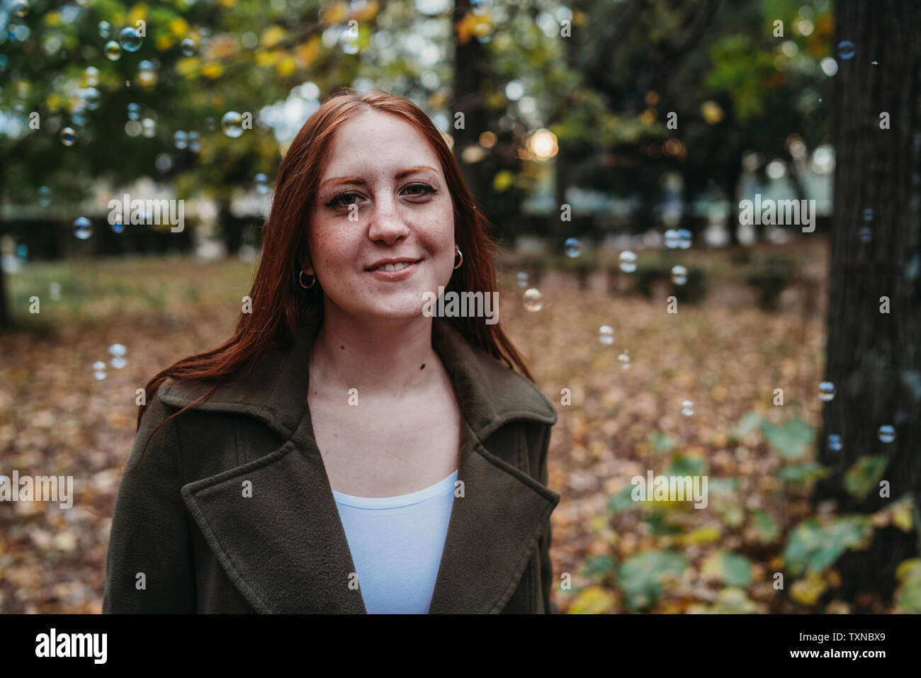 Young woman with long red hair amongst floating bubbles in autumn park, head and shoulder portrait Stock Photo