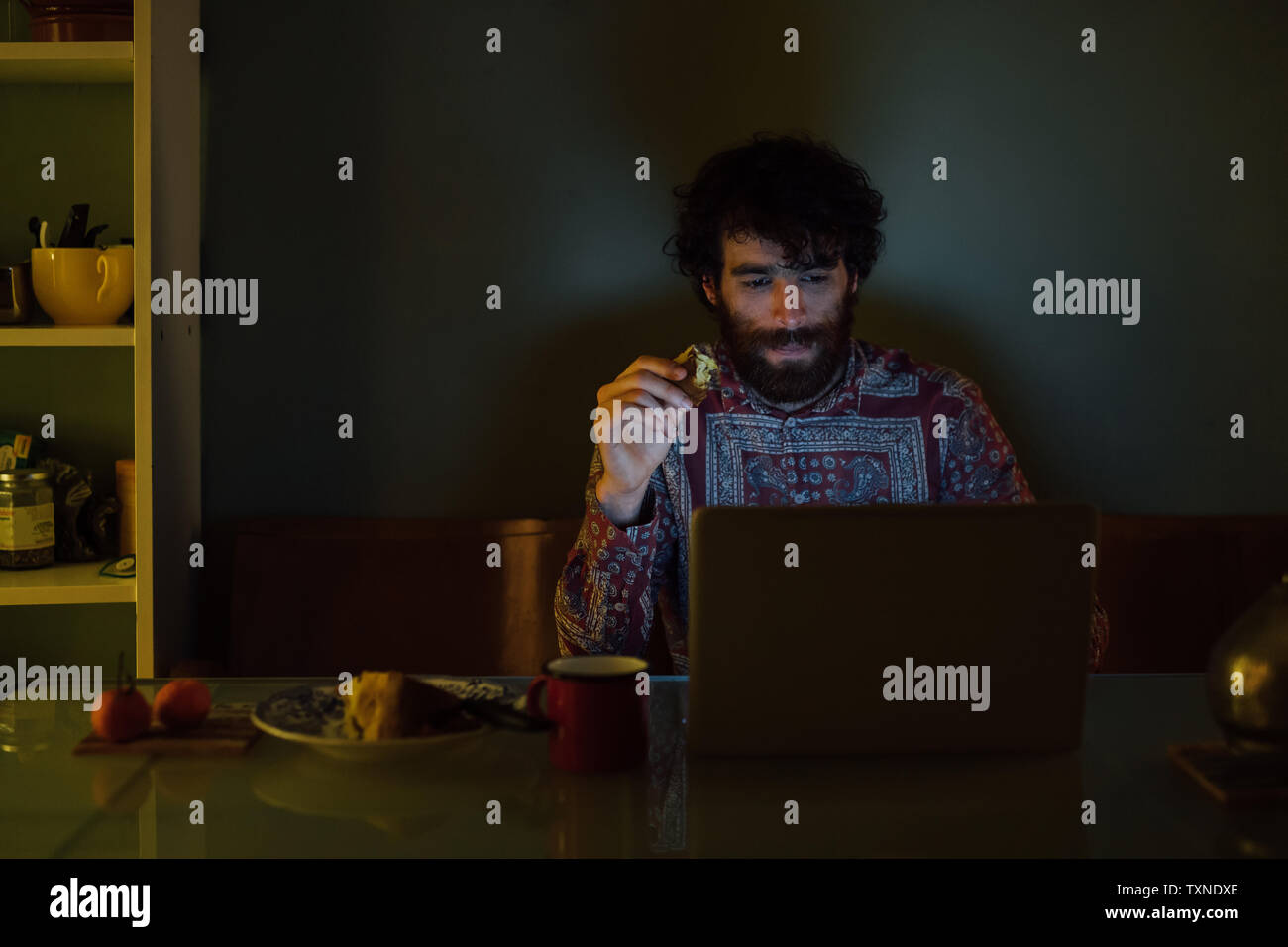 Bearded young man having breakfast, using laptop at home Stock Photo