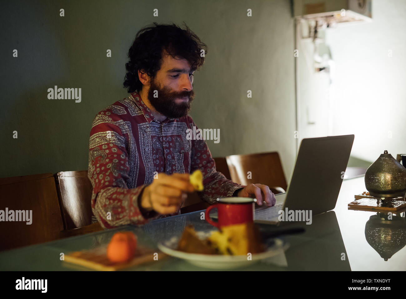 Bearded young man having breakfast, using laptop at home Stock Photo