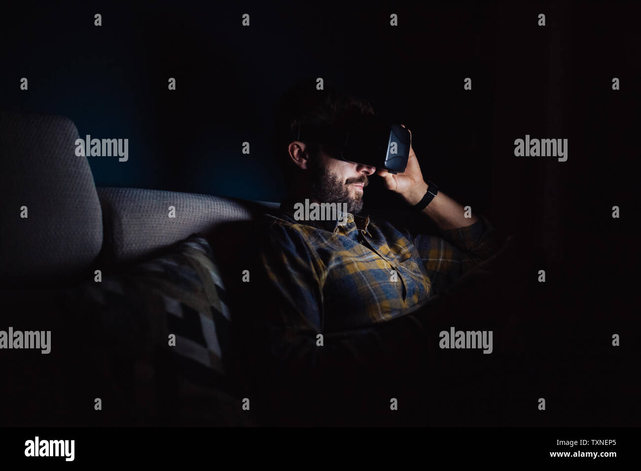 Bearded mid adult man sitting on sofa in darkness looking down through virtual reality headset Stock Photo