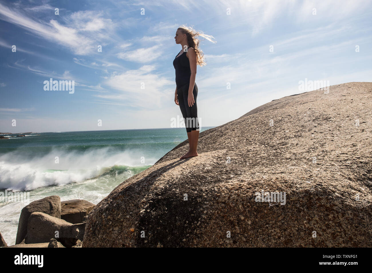 Young woman practicing yoga taking a break looking out from beach boulder, Cape Town, Western Cape, South Africa Stock Photo