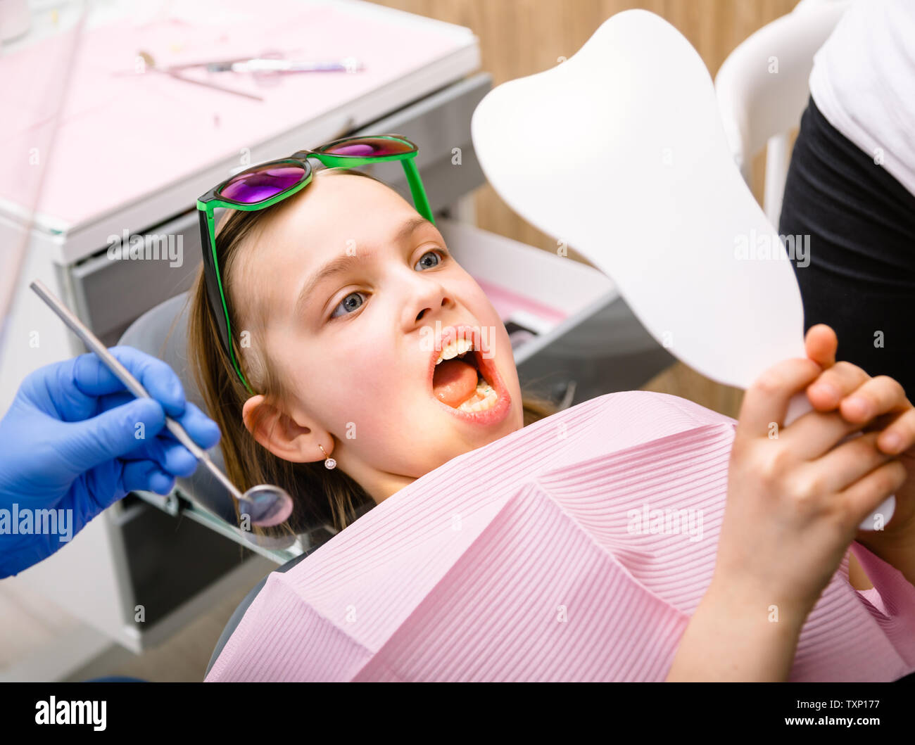 Preteen girl sitting in a dental chair inspecting repaired teeth looking at tooth-shaped mirror in pediatric dental clinic Stock Photo