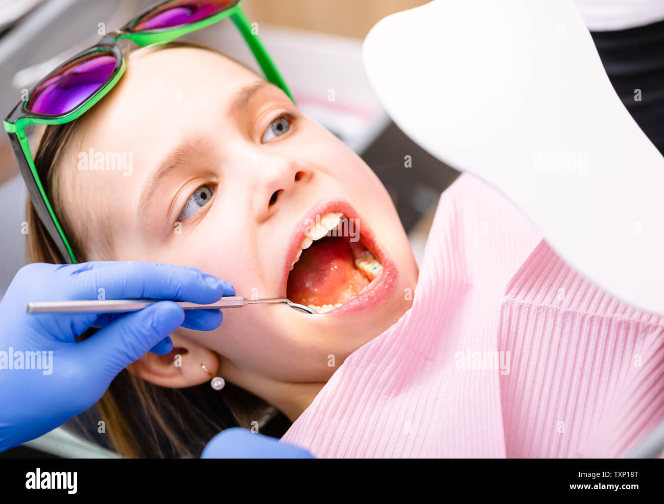 Preteen girl sitting in a dental chair inspecting repaired teeth looking at tooth-shaped mirror in pediatric dental clinic Stock Photo