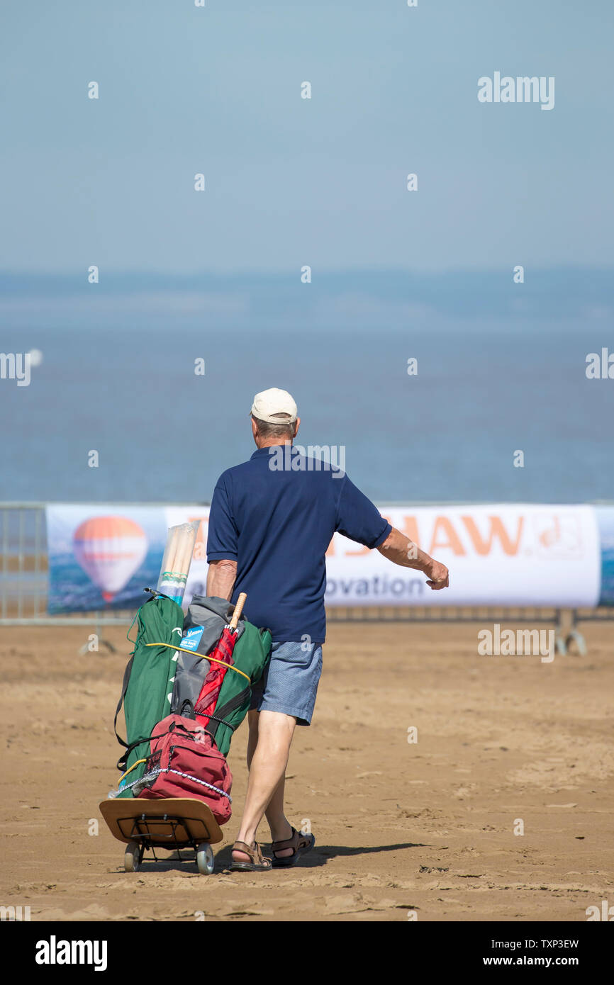 Man in summer shorts, open sandals & sports cap, pulling beach trolley along the sand, Weston-super-Mare, UK. Father fetching beach equipment. Stock Photo