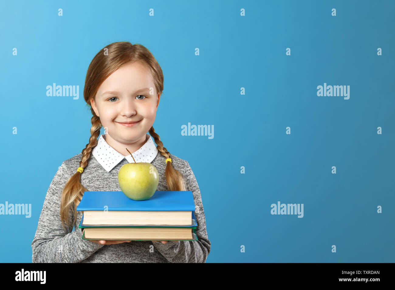 Portrait of a cheerful little girl on a blue background. Schoolgirl is holding a stack of books and an apple. Back to school. The concept of education Stock Photo