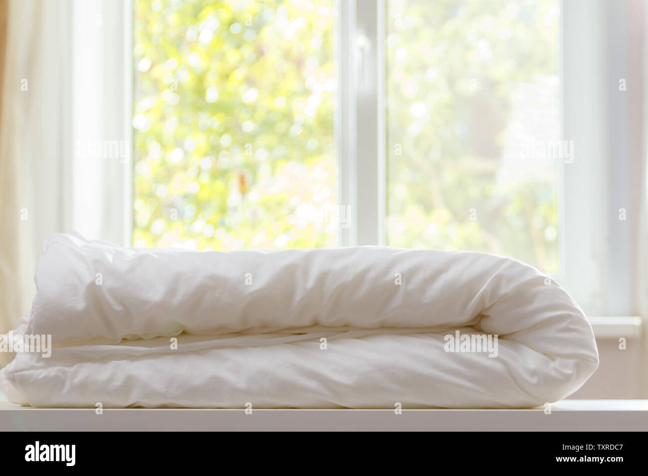 A white duvet is lying on the dresser against the blurred background of the window. Stock Photo