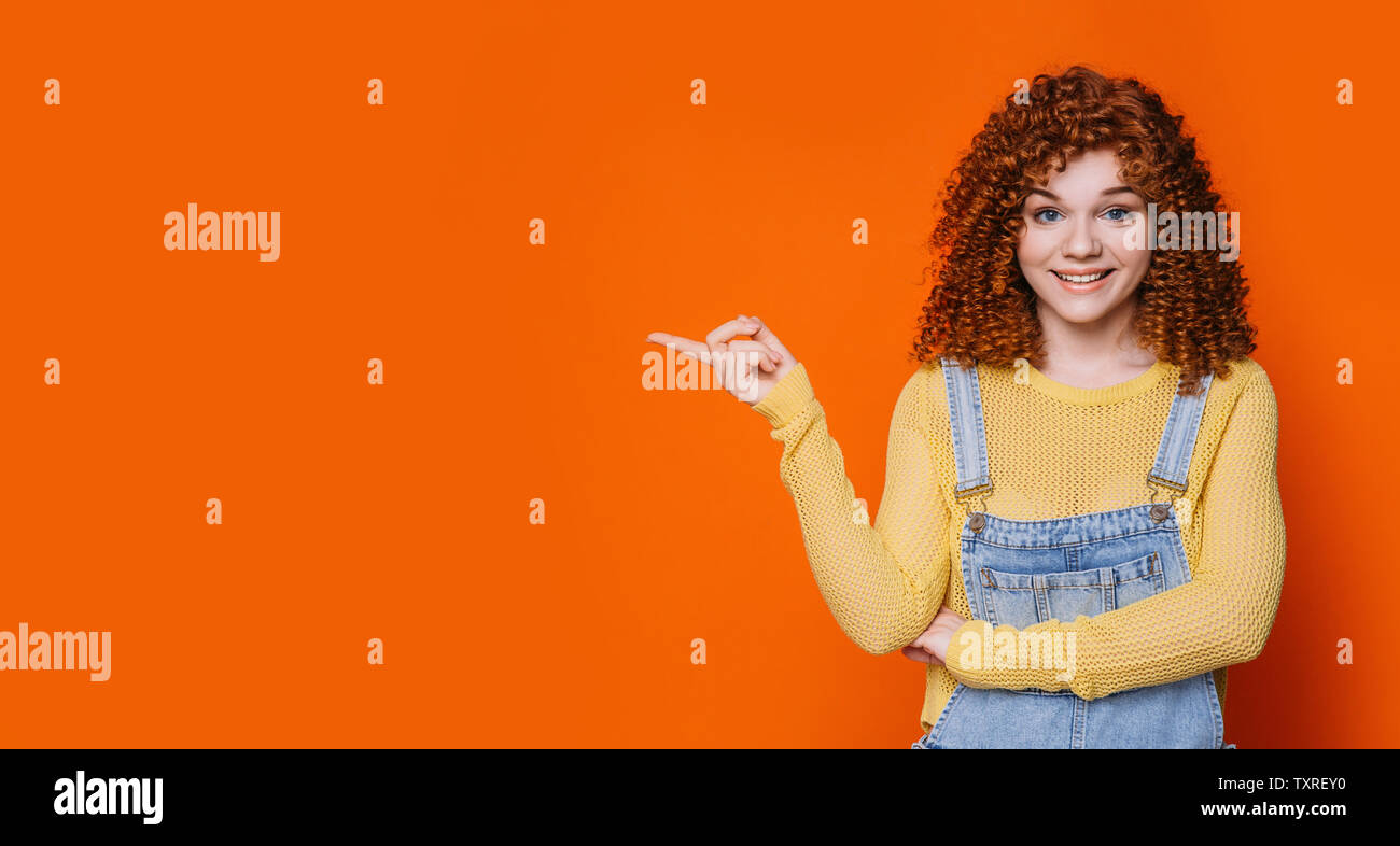 curly red-haired woman smiling and pointing hands into empty orange background.Hey you look this Stock Photo