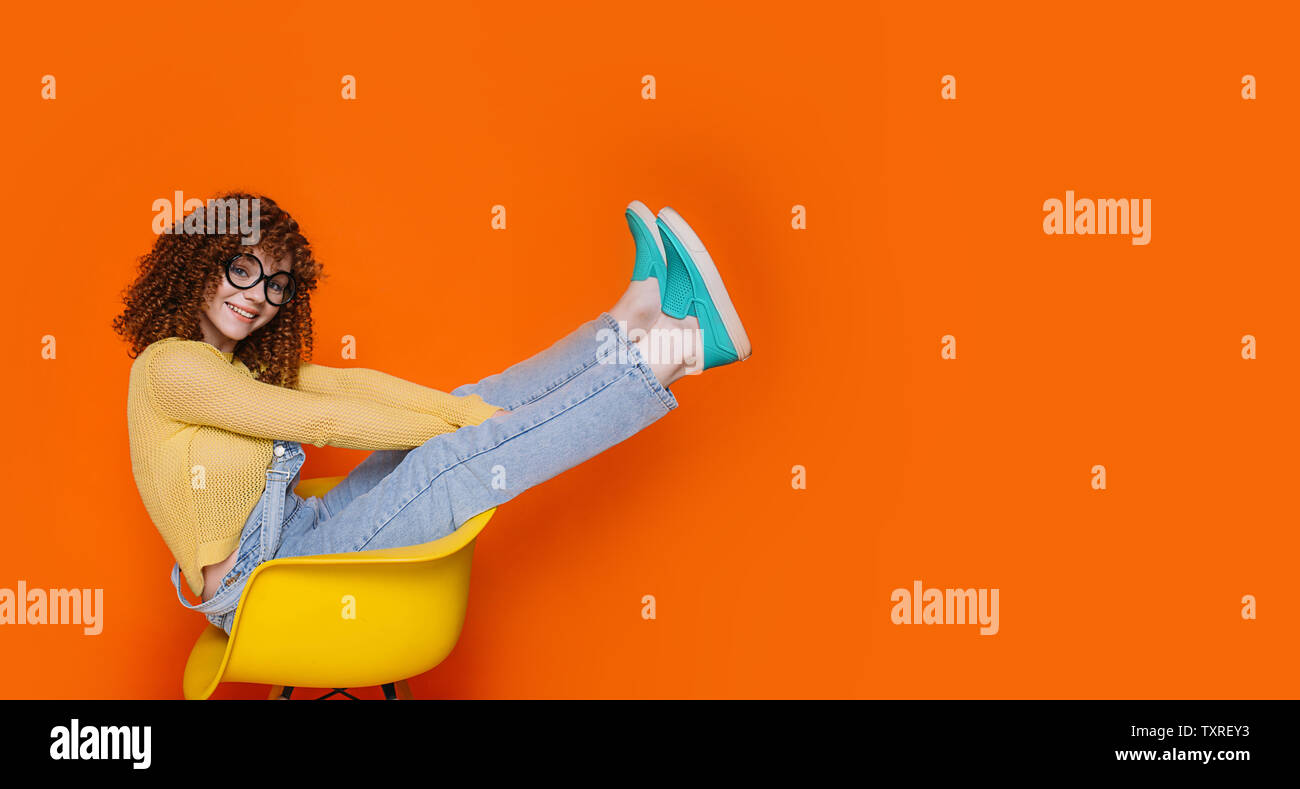 Trendy teenage with curly hair wearing denim overalls and yellow jacket sitting on chair with raised legs. funny young woman on orange background Stock Photo