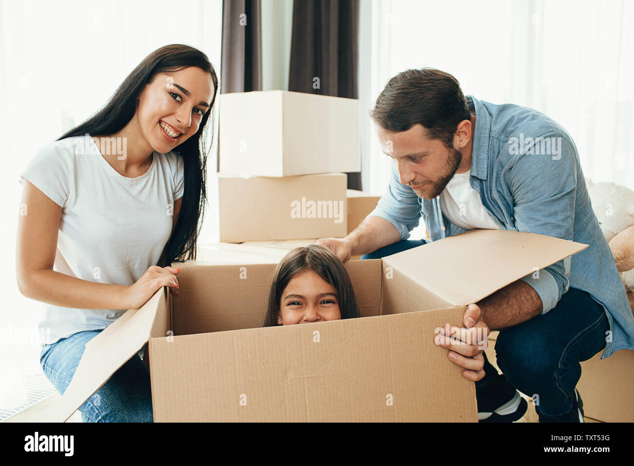 Happy family having fun while moving into new apartment. Mother, father and daughter together smiling, with Cardboard boxes on background Stock Photo