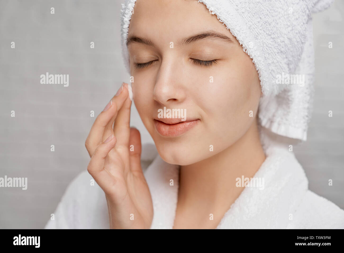 Close up of young, attractive woman with closed eyes applying moisturizer on face. Pretty girl with perfect skin in white bathrobe with white towel posing on grey background. Stock Photo
