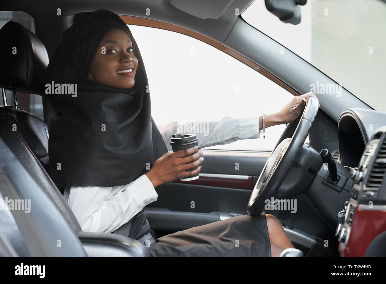Cheerful african woman holding hand on steering wheel and coffee cup while driving car. Beautiful young muslim woman in black hijab sitting in car, looking at camera. Stock Photo
