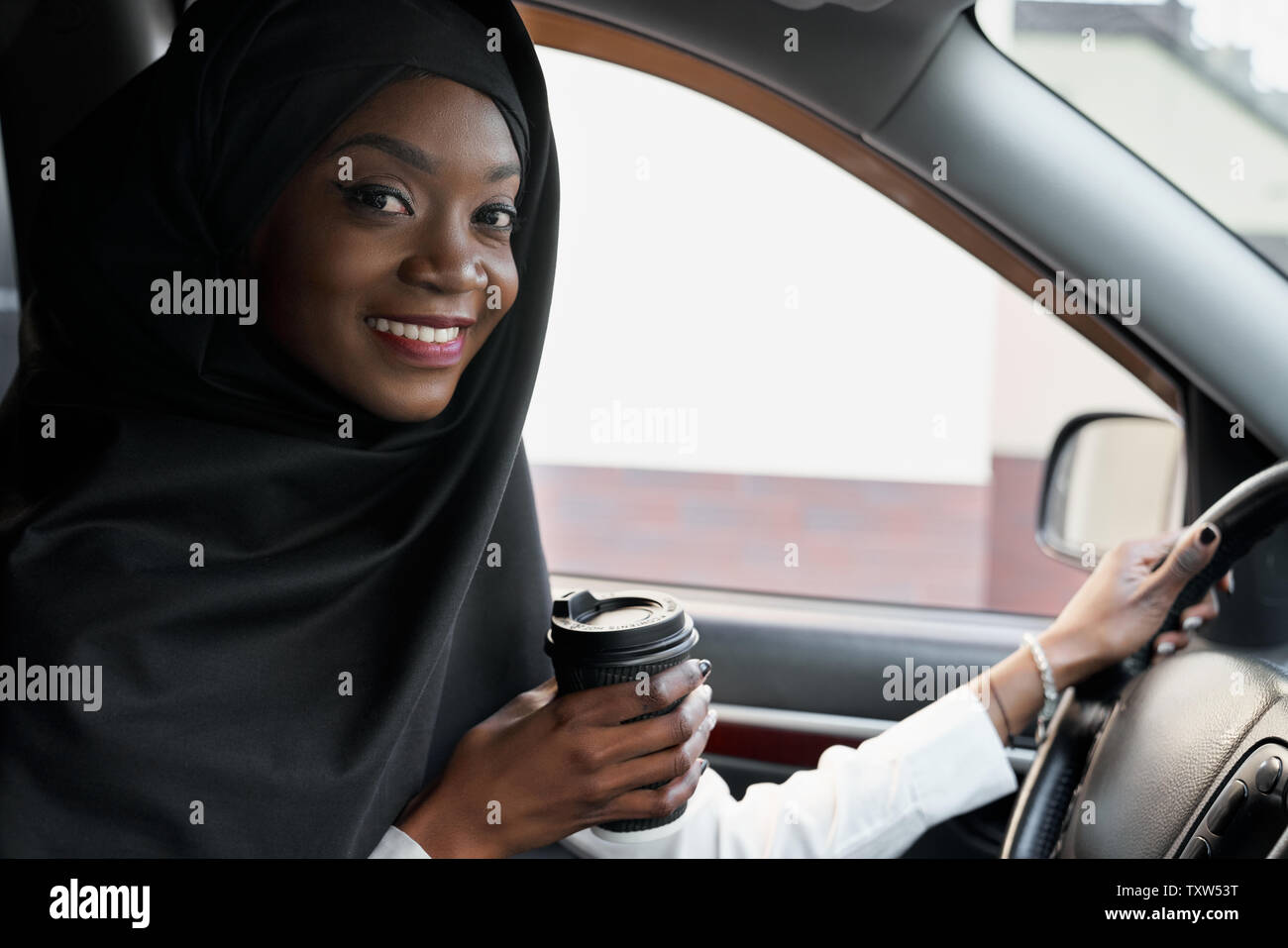 Positive, cheerful african girl sitting in car posing, holding hand on steering wheel. Beautiful muslim woman wearing in black hijab with perfect smile looking at camera, holding coffee cup. Stock Photo