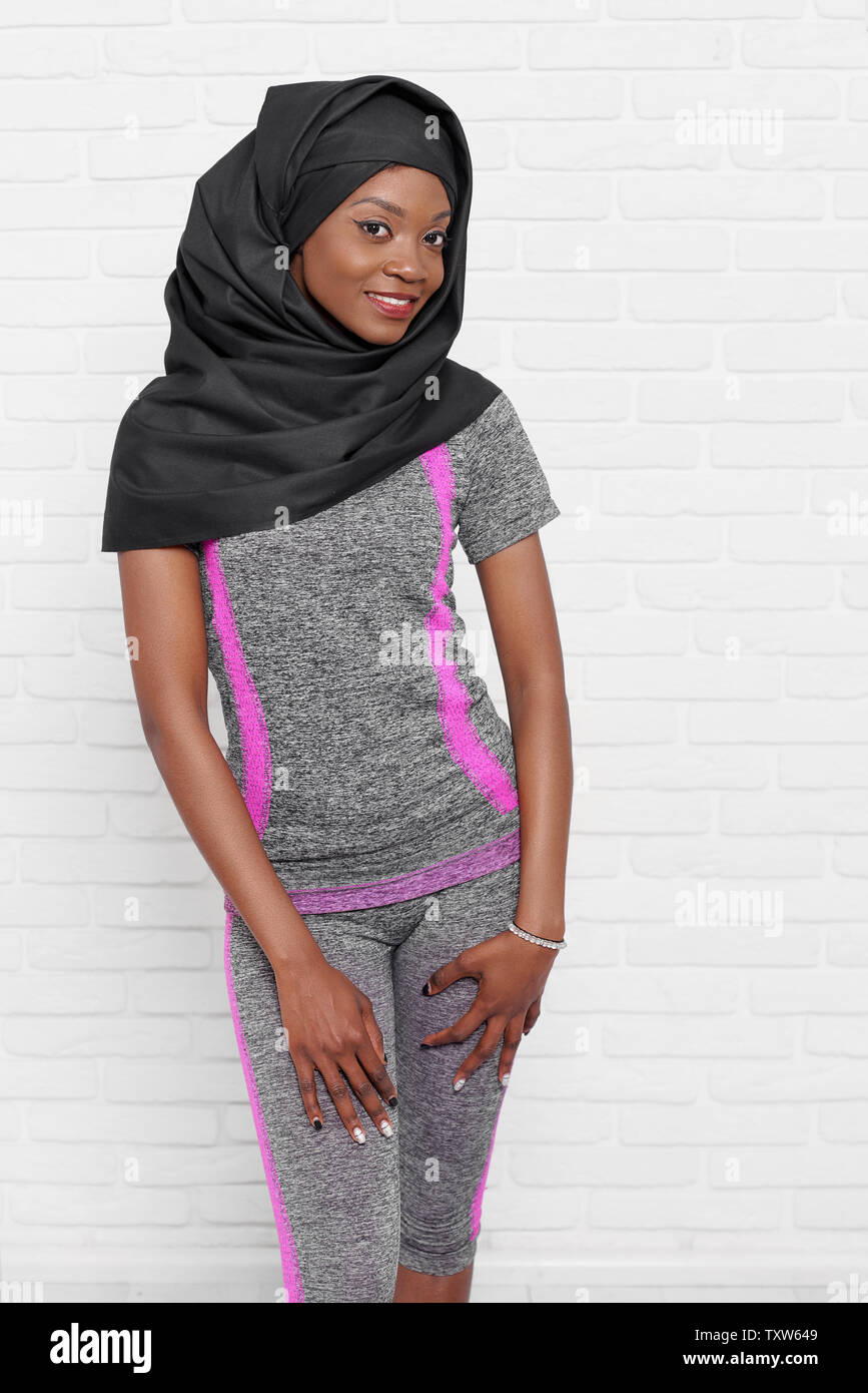 Stylish young muslim woman wearing in black hijab and grey sportswear  posing against white brick wall. Beautiful, positive, slim african model  standing, looking at camera, smiling Stock Photo - Alamy