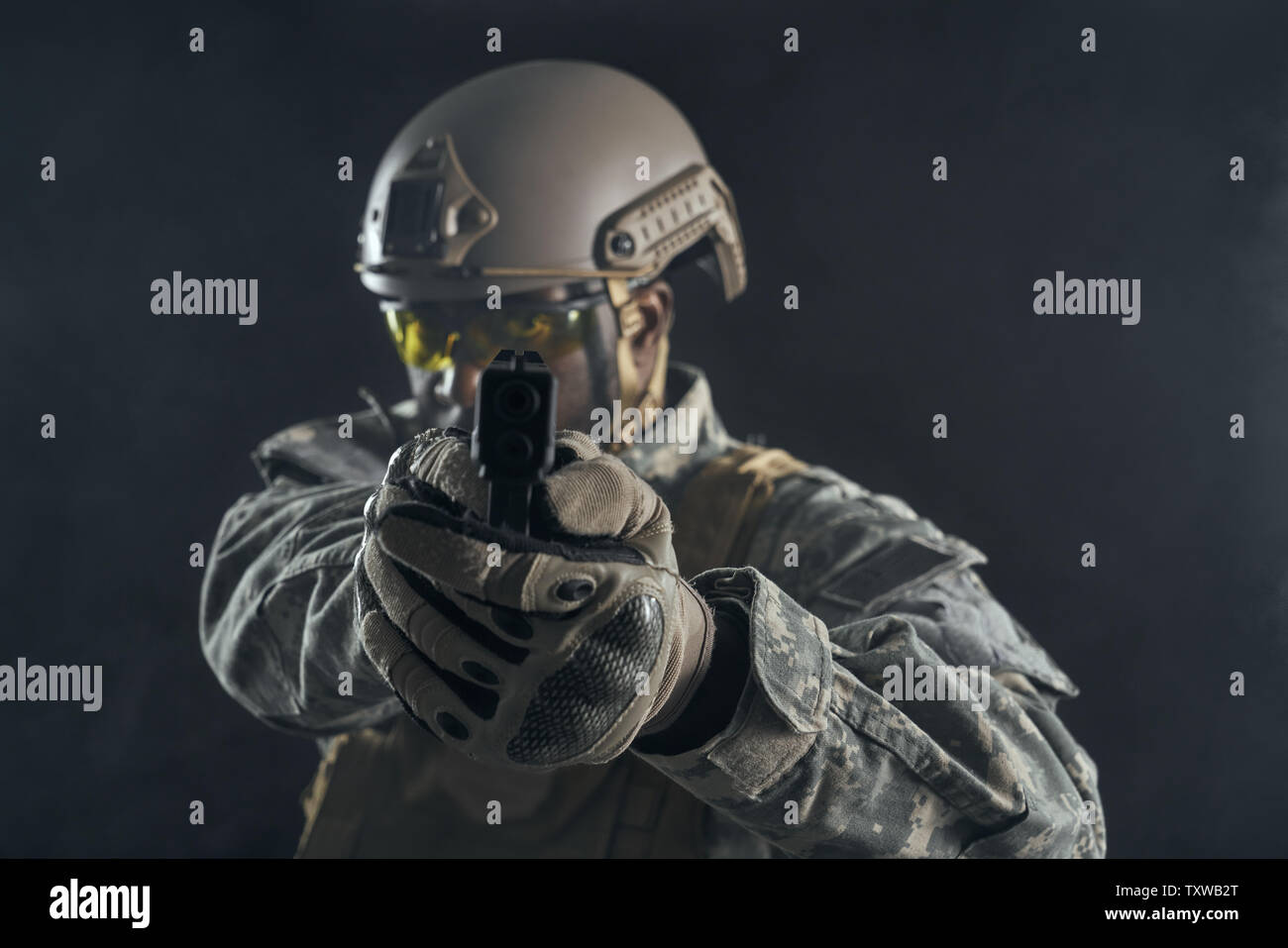Front view of seriously American soldier in uniform holding modern gun in hands and shooting at camera. Brave ranker wearing helmet keeping position, posing in darkness. Black background. Stock Photo